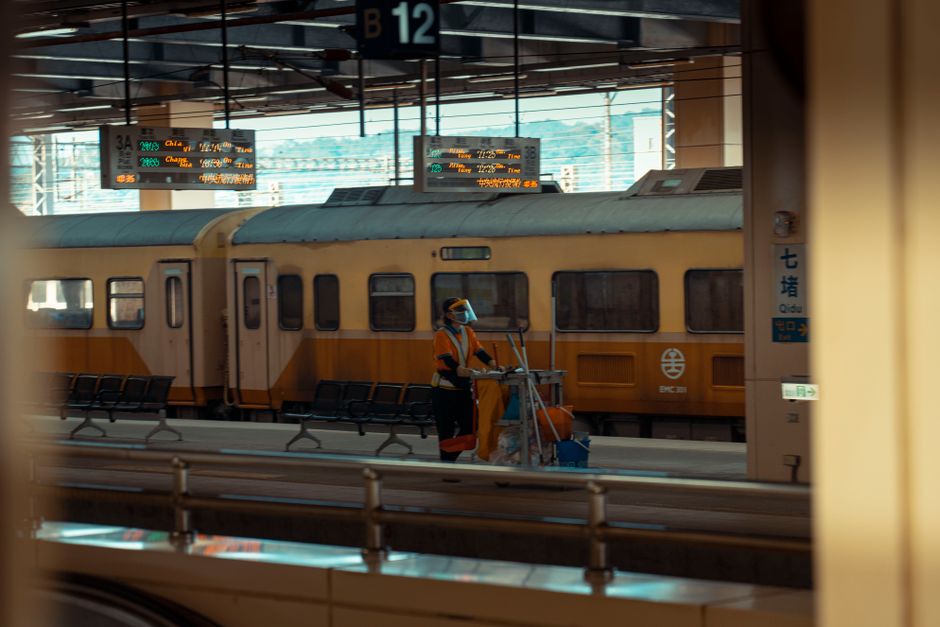 A worker wearing a high-visibility vest and black clothing stands on a train station platform next to a large cart filled with black, gray, and orange garbage bags, along with some miscellaneous items
